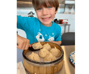 smiliing boy in blue shirt about to eat a soup dumpling from tiger soup dumplings